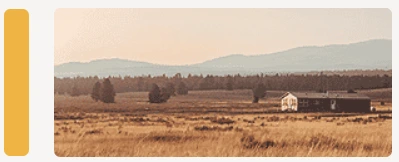 A peaceful rural landscape featuring a small white farmhouse and barn set in a golden field of dry grass, with a backdrop of dense trees and distant blue mountains under a soft, warm sky.